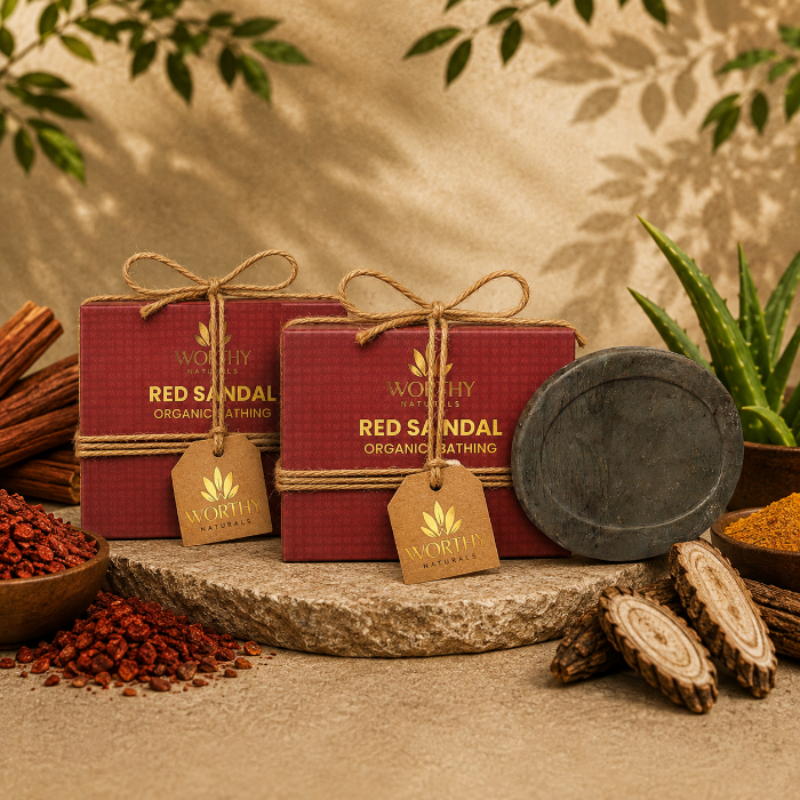 Two red rectangular boxes of Worthy of Kerala Red Sandal Organic Bathing tied with twine and hanging tags, displayed on a stone slab with bath ingredients and a dark round stone vessel in a styled setting.